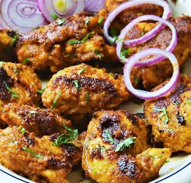 Achari Murg Tikka (300 g) : Morceaux de poulet épicés, dorés et grillés, à la texture croustillante et carbonisée, garnis d'herbes hachées et servis avec des oignons rouges émincés.