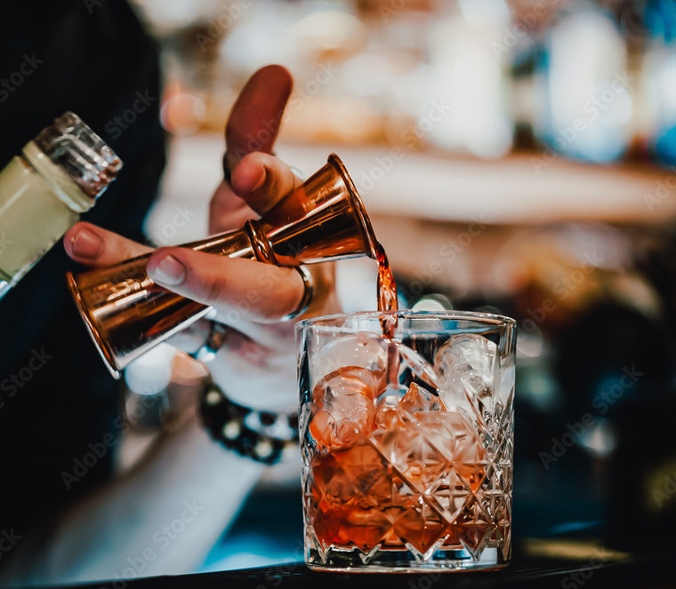 Un barman verse OPEN BAR VINS & CHAMPAGNES dans un verre rempli de glaçons, avec un bar flou en arrière-plan.