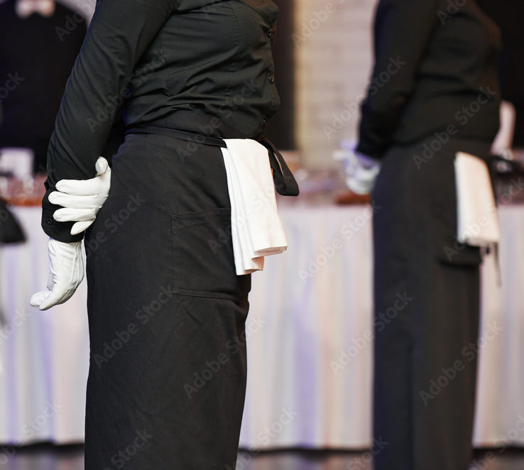 Deux serveurs en uniforme noir, gants blancs et serviettes à la taille se tiennent formellement devant une table recouverte d'une nappe blanche, prêts à servir lors de l'événement OPEN BAR VINS & CHAMPAGNES (Ne payez que ce que vous consommez).