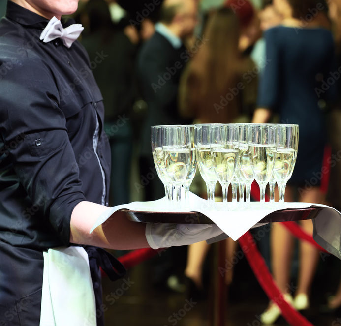 Lors d'un événement formel, un serveur en chemise noire et nœud papillon blanc sert OPEN BAR VINS & CHAMPAGNES (Ne payez que ce que vous consommerez) sur un plateau de coupes de champagne, avec des invités flous et une corde rouge en arrière-plan.