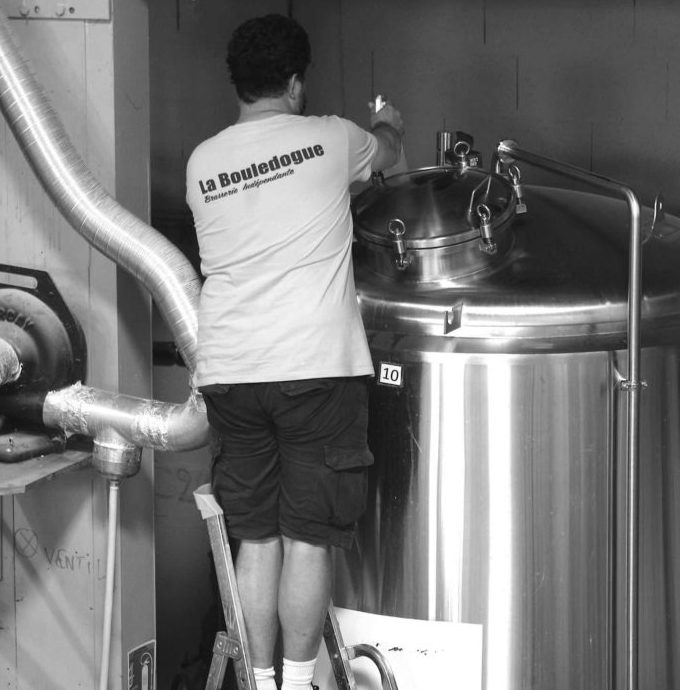 Une personne sur un escabeau inspecte une grande cuve en inox dans une brasserie. Le dos de leur chemise porte l'inscription "La Bouledogue", en référence à la FÛT DE BIÈRE BLONDE 5° 30L (Bière artisanale bouledogue).