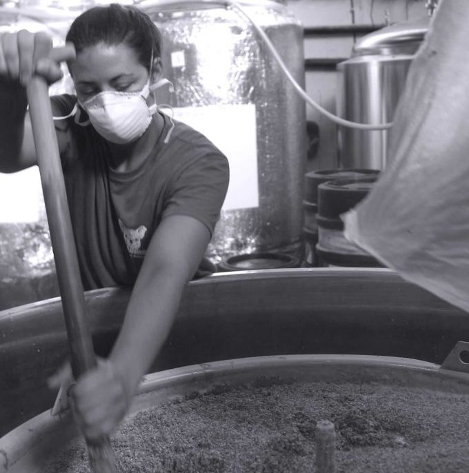 Une personne masquée remue un grand récipient de FÛT DE BIÈRE BLONDE 5° 30L (Bière artisanale bouledogue) dans une brasserie industrielle.