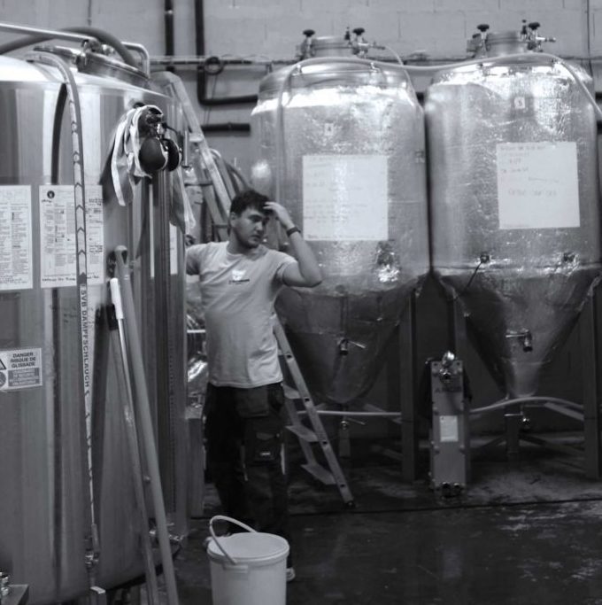 Un homme se tient la tête dans une brasserie, à côté de grandes cuves de fermentation en métal. On aperçoit du matériel de brassage et une échelle. L'image en noir et blanc représente la FÛT DE BIÈRE BLONDE 5° 30L (Bière artisanale bouledogue) dans un environnement industriel.