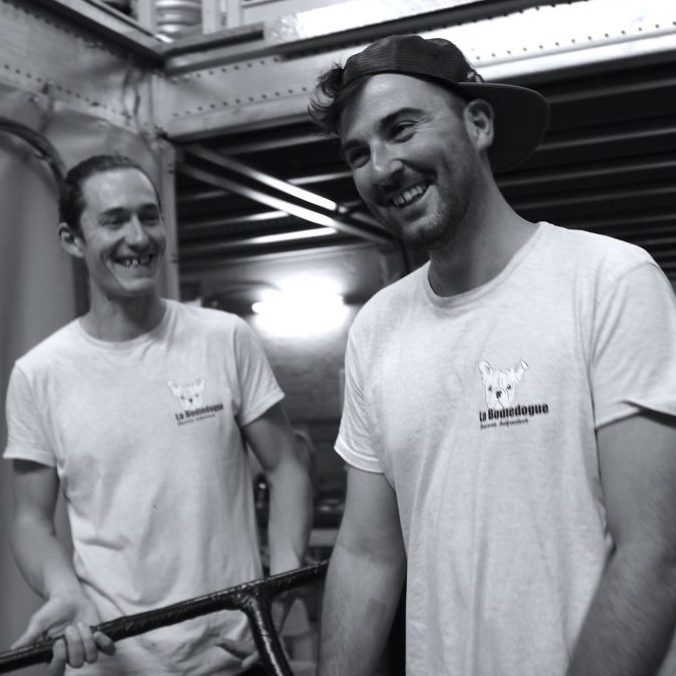 Deux jeunes hommes en t-shirt blanc sourient à l'intérieur sous un éclairage industriel, l'un tenant une barre métallique. Ils ont l'air détendu à côté d'un FÛT DE BIÈRE BLONDE 5° 30L (Bière artisanale bouledogue). La photo est en noir et blanc.