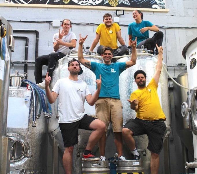 Six joyeux lurons aux chemises colorées posent devant des cuves de brassage, certains debout sur des fûts, célébrant la FÛT DE BIÈRE BLONDE 5° 30L (Bière artisanale bouledogue) dans le cadre d'une brasserie industrielle.