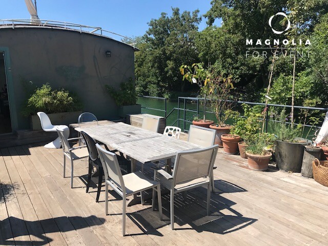 Terrasse extérieure en bois avec une longue table et des chaises dépareillées, entourée de plantes en pot et bordée d'arbres et d'une balustrade en métal, mettant en vedette LA PÉNICHE DE JEROME (92040). Le logo Magnolia For Event se trouve dans le coin supérieur droit.