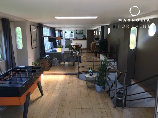 Espace de vie spacieux et moderne avec sols en bois, baby-foot, plantes, canapés près de grandes fenêtres rondes. L'escalier descend. Le logo de LA PÉNICHE DE JEROME (92040) apparaît dans le coin supérieur droit.