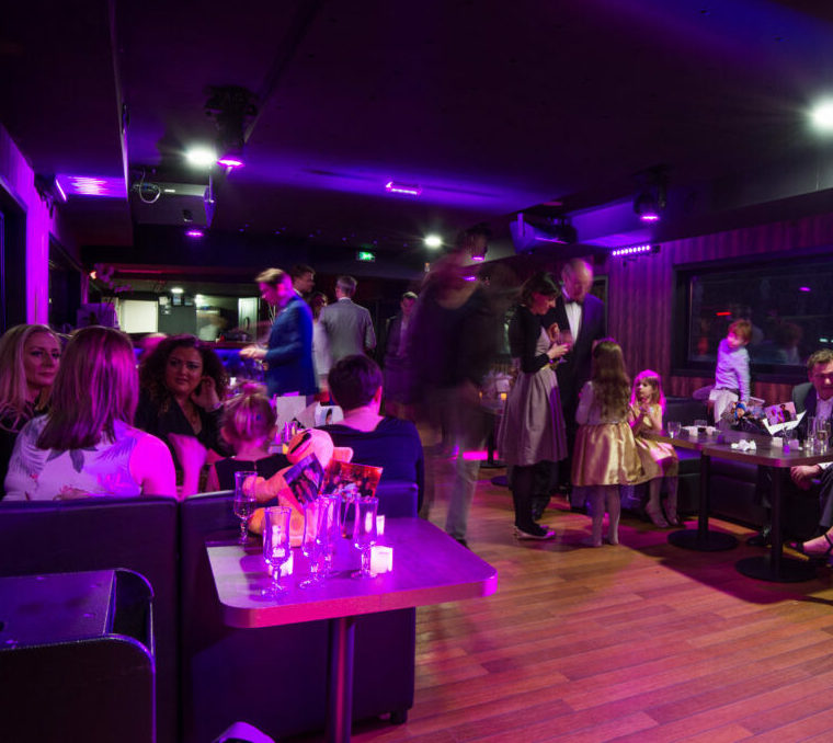 À LA BELLE PÉNICHE (75005), adultes et enfants se retrouvent dans un salon faiblement éclairé par des lumières violettes, discutant et dégustant des boissons à des tables ou debout dans une atmosphère vivante et sociale.