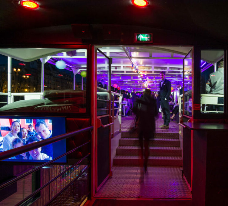 Une personne monte des marches illuminées dans LA BELLE PÉNICHE (75005), une salle moderne aux lumières violettes et rouges, où les invités sont assis, où un membre du personnel les accueille à l'entrée et où une photo de famille est affichée sur un écran à gauche.