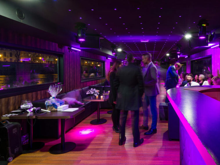 Un groupe animé socialise à LA BELLE PÉNICHE (75005), un salon moderne à l'éclairage violet et aux grandes fenêtres offrant une vue nocturne sur la ville, tandis que les invités se mêlent aux tables ou se détendent dans des sièges confortables.