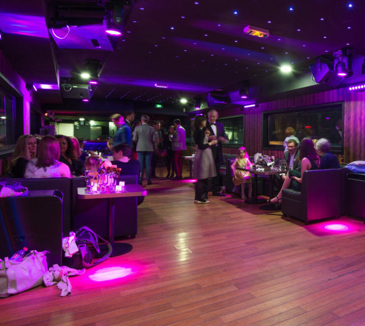 À LA BELLE PÉNICHE (75005), des groupes discutent à des tables tandis que d'autres se mêlent à un salon animé. L'éclairage tamisé aux teintes violettes et le plancher en bois reflètent l'ambiance vibrante et colorée du lieu.