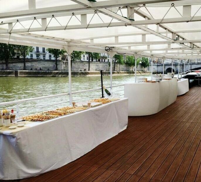 Une table de buffet drapée de blanc est dressée sur la terrasse en bois de LA BELLE PÉNICHE (75005), au bord de la rivière, avec un auvent au-dessus de la tête et une vue sur les bâtiments et les arbres de l'autre côté de l'eau.