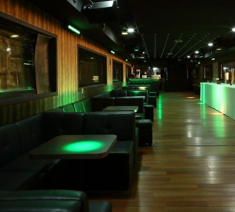LA BELLE PÉNICHE (75005) propose un salon moderne avec des cabines en cuir sombre, des planchers en bois, des murs et un plafond éclairés par des LED vertes, de grandes fenêtres d'un côté, et un bar élégant en face. L'espace est actuellement vide.