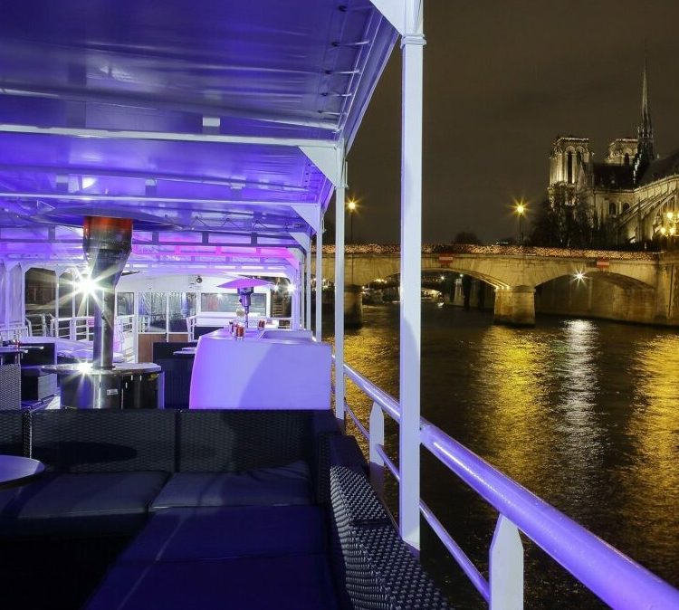 Découvrez les vues depuis LA BELLE PÉNICHE (75005) - un bateau moderne à l'éclairage violet qui navigue sur la Seine la nuit, mettant en valeur la cathédrale Notre-Dame illuminée et un pont voisin se reflétant sur l'eau.