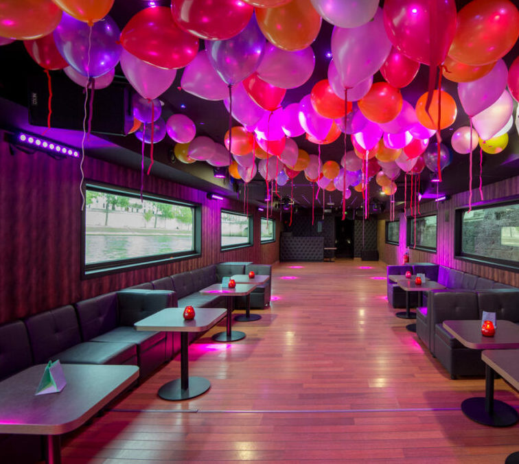 LA BELLE PÉNICHE (75005) propose un espace festif avec des ballons colorés au plafond, un éclairage violet, des canapés noirs vides, des petites tables, de grandes fenêtres murales et un sol en bois chaleureux pour votre fête décorée.