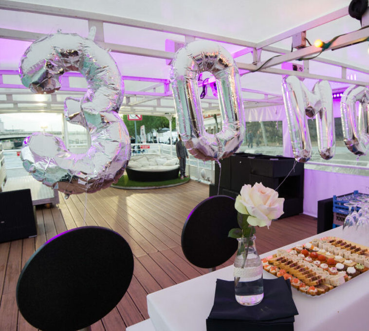 À LA BELLE PÉNICHE (75005), des ballons argentés de 30 mètres de diamètre sont mis en valeur dans une salle décorée d'un éclairage violet, tandis qu'une rose blanche dans un vase et des desserts assortis ornent la table au premier plan.