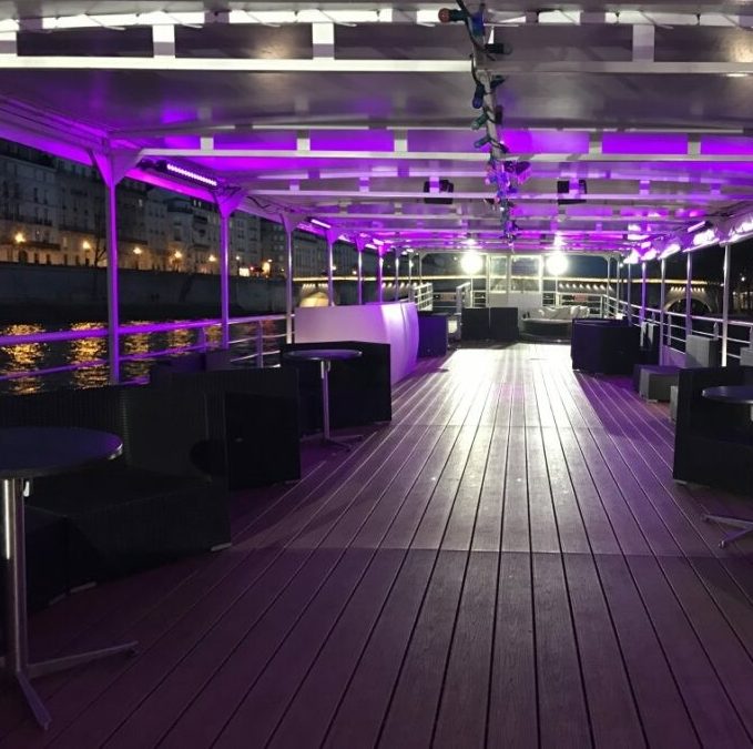 Sur LA BELLE PÉNICHE (75005), une terrasse en plein air avec un sol en bois est équipée de chaises et de tables noires modernes sur les côtés, éclairées par des lumières violettes. Les lumières de la ville et les bâtiments brillent sur l'eau en arrière-plan.