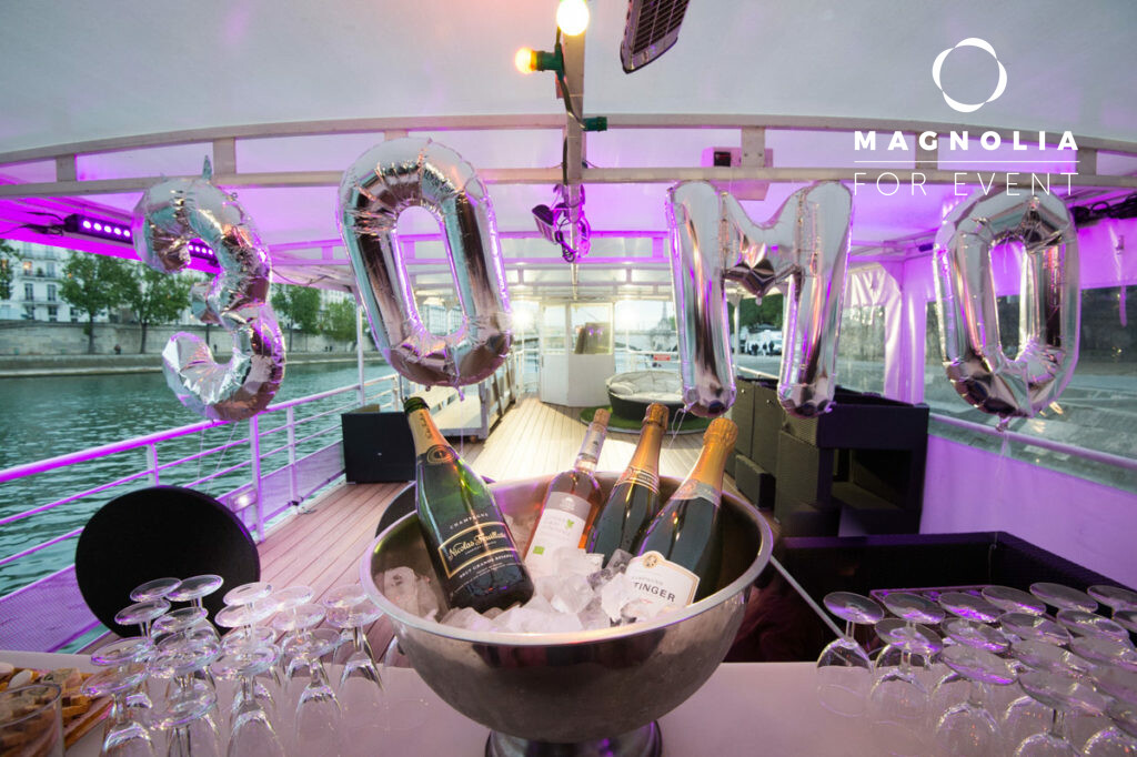 LA BELLE PÉNICHE (75005) : Un bateau décoré avec des ballons argentés "30MO", des bouteilles de champagne dans un seau à glace, des verres sur une table, un éclairage violet, un paysage urbain en toile de fond et le logo Magnolia For Event.