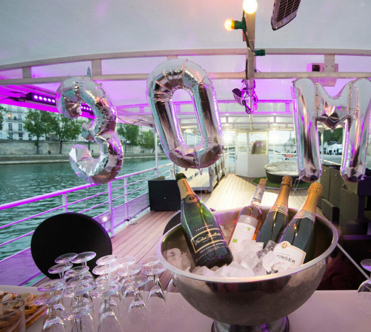 Des ballons métalliques portant l'inscription "30M" ornent un bateau LA BELLE PÉNICHE (75005), avec du champagne sur de la glace, des verres à proximité, le tout sous des lumières violettes ; un fleuve et un paysage urbain forment l'arrière-plan.