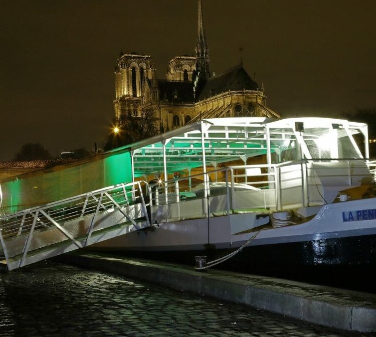 LA BELLE PÉNICHE (75005) est un bateau fluvial brillamment éclairé, amarré sur la Seine la nuit, avec la cathédrale Notre-Dame illuminée en arrière-plan et les pavés mouillés reflétant les lumières vibrantes.