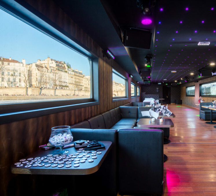 LA BELLE PÉNICHE (75005) dispose d'un salon moderne avec de grandes fenêtres donnant sur les bâtiments de la ville de l'autre côté d'une rivière, des canapés gris, des planchers en bois, des tables avec des boutons et des plafonniers colorés éclairés par la lumière du jour.
