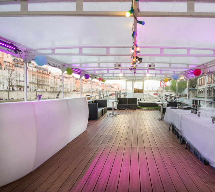 LA BELLE PÉNICHE (75005) propose un pont de bateaux en plein air avec des planchers en bois, des guirlandes lumineuses et des ballons, des sièges de salon, un bar sur la gauche et des tables pour prendre un verre sur la droite, le tout amarré au bord de l'eau de la ville.
