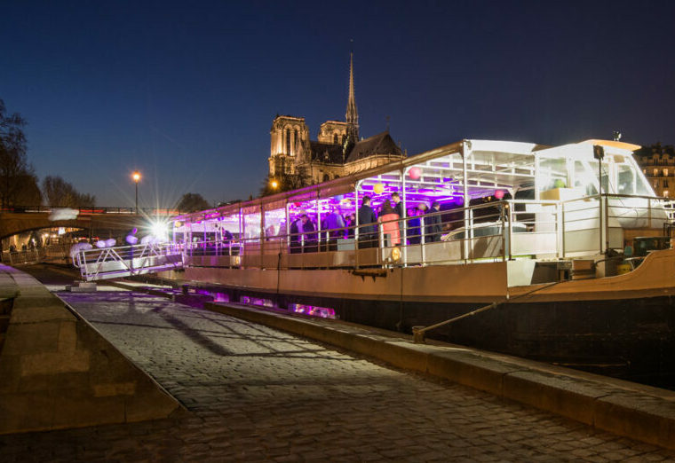 Le LA BELLE PÉNICHE (75005), illuminé de lumières intérieures violettes, est amarré de nuit sur un quai pavé, la cathédrale Notre-Dame étant visible en arrière-plan sous un ciel dégagé.