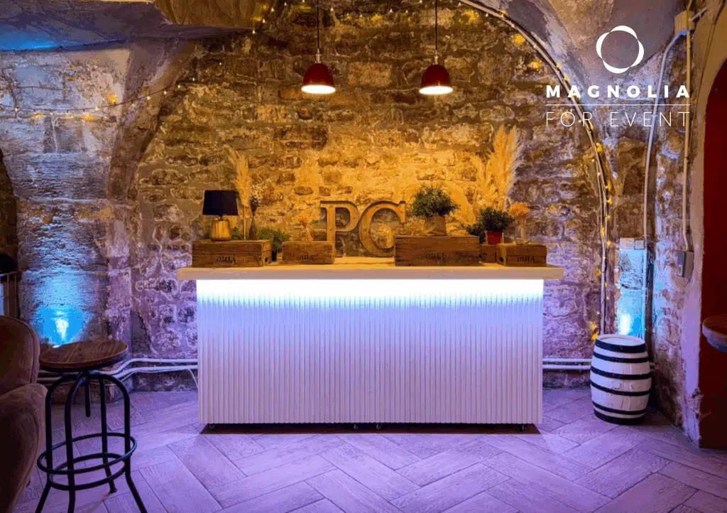 LE PETIT PARIS CACHÉ (75004) dispose d'un bar rustique aux murs de pierre, d'un éclairage chaleureux, de deux lampes rouges suspendues, d'un comptoir blanc brillant, de plantes décoratives et du logo Magnolia For Event dans le coin supérieur droit.