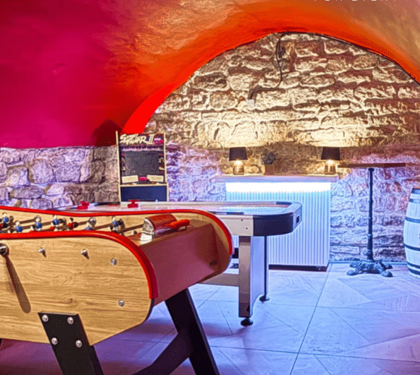 LE PETIT PARIS CACHÉ (75004) dispose d'une salle de jeux confortable avec un mur en pierre, un plafond voûté rouge, des tables de baby-foot et d'air hockey, une machine à sous, un décor de tonneaux et un éclairage chaleureux provenant de deux lampes sur l'étagère du fond.
