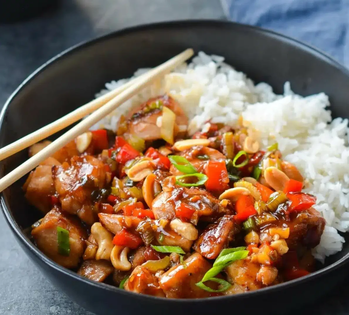 Un bol noir contient le POULET IMPÉRIAL (250 g/pers) : riz blanc, poulet sauté, poivrons rouges, cacahuètes, oignons verts hachés et sauce, avec des baguettes posées sur le dessus.