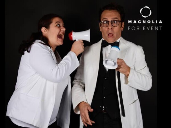 Une femme en veste blanche crie dans un mégaphone à l'adresse d'un homme surpris en blanc, tous deux tenant des mégaphones bleus, sur un fond noir avec le logo DUO DE FAUX SERVEURS MAGICIENS.