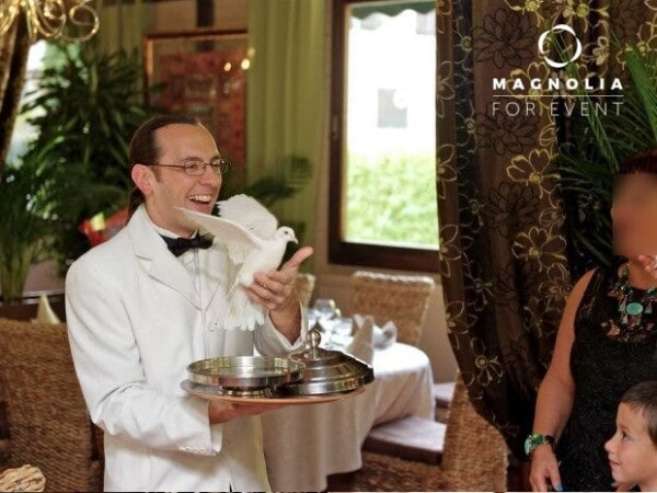 Un homme souriant en costume blanc fait un tour de magie avec un plateau en argent et une colombe pour des enfants lors d'un événement en intérieur, entouré de plantes et de rideaux. Le logo Magnolia For Event apparaît, avec le DUO DE FAUX SERVEURS MAGICIENS.