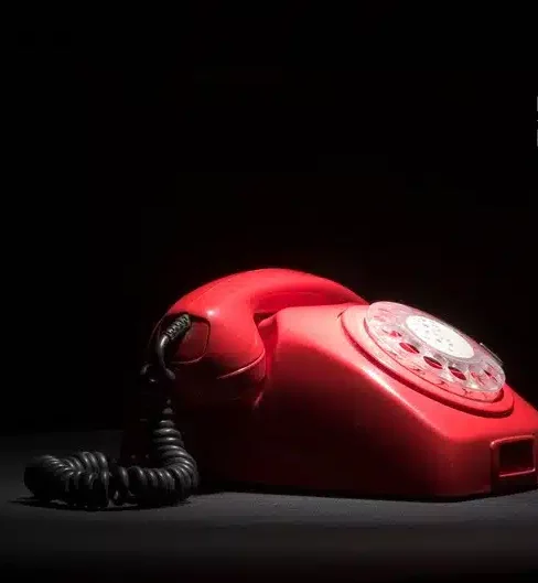 Le LIVRE D'OR AUDIO, un téléphone vintage à cadran rotatif rouge avec un cordon noir, est posé sur une surface sombre sur fond noir, partiellement éclairé par la gauche.