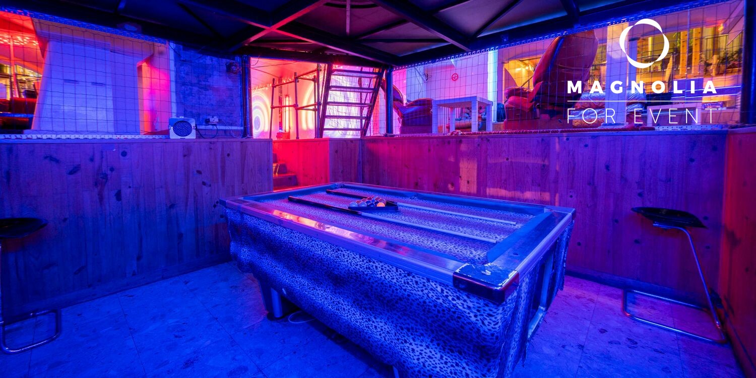 LE LOFT DE TITO (75011) : Comprend un billard imprimé léopard dans une salle en bois avec des néons, des tabourets de bar, un panneau "Magnolia For Event" et un escalier menant à des places assises supplémentaires à l'arrière-plan.