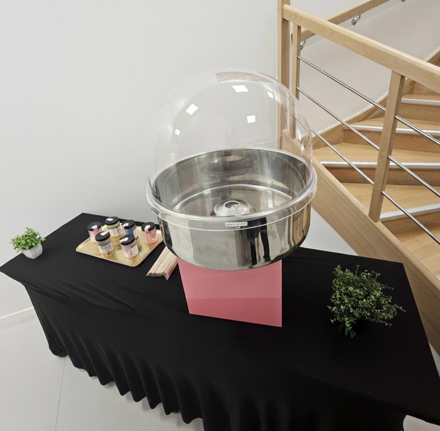 La machine à barbe à papa ANIMATION BARBE A PAPA avec un dôme transparent et un socle rose est présentée sur une nappe noire, accompagnée de pots de sucre coloré, de bâtons en bois, de deux petites plantes en pot, à côté d'un escalier en bois.