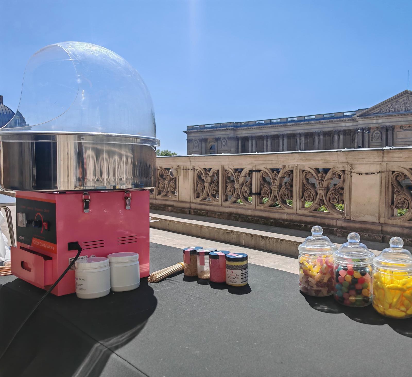 La machine à barbe à papa ANIMATION BARBE A PAPA est présentée en plein air sur une table, entourée de bocaux de bonbons colorés, avec des bâtiments historiques en pierre et un ciel bleu clair en arrière-plan.