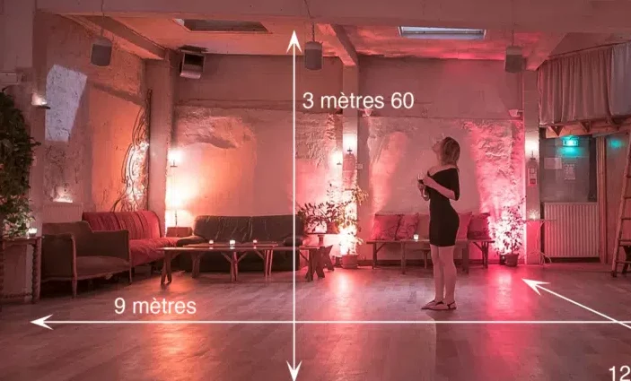 Une femme en robe noire se tient dans LE LOFT DES BATIGNOLES (75017), chaleureusement éclairé, avec des murs apparents, des canapés et des plantes. Des lignes blanches et un texte en français indiquent les dimensions de la pièce : 9 mètres sur 3,6 mètres.