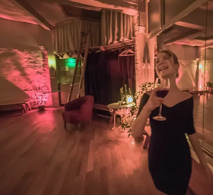 Une femme en robe noire sourit en tenant un verre de vin rouge dans LE LOFT DES BATIGNOLES (75017), légèrement éclairé, avec des planchers en bois, un fauteuil rouge, des plantes et un espace scénique à l'arrière-plan.