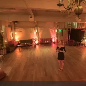 Une femme en robe noire et sandales se tient seule dans LE LOFT DES BATIGNOLES (75017), une pièce spacieuse et faiblement éclairée avec des planchers en bois, des lumières chaudes, des canapés et des bougies qui créent une atmosphère intime et confortable.