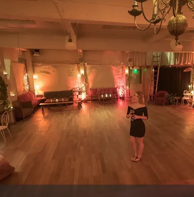 Une femme en robe noire et sandales se tient seule dans LE LOFT DES BATIGNOLES (75017), une pièce spacieuse et faiblement éclairée avec des planchers en bois, des lumières chaudes, des canapés et des bougies qui créent une atmosphère intime et confortable.