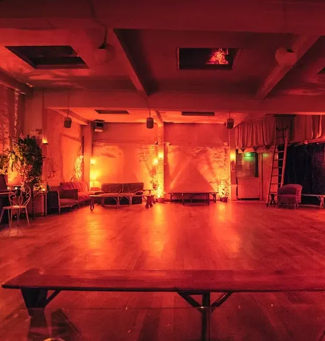 LE LOFT DES BATIGNOLES (75017) dispose d'une salle spacieuse et peu éclairée, avec un éclairage rouge, des sols en bois, des canapés le long des murs, des plantes en pot, des lampes d'ambiance et une banquette vide pour une ambiance intime et chaleureuse.