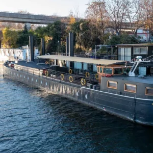 Le LE JAZZ HARMONY (75011) est un grand bateau noir avec des bouées de sauvetage jaunes, de hautes cheminées, plusieurs fenêtres et un pont supérieur, amarré sur une rivière près d'arbres et d'un viaduc.