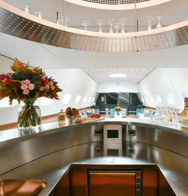 Le LE JAZZ HARMONY (75011) présente un intérieur d'avion luxueux avec un bar moderne, des fleurs fraîches, des boissons, des en-cas sur un comptoir brillant, des sièges de salon élégants et de grandes fenêtres en arrière-plan.