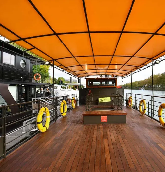 LE JAZZ HARMONY (75011) présente un pont de bateau couvert avec un toit jaune-orange vif, un plancher en bois, des rambardes de sécurité et plusieurs bouées de sauvetage jaunes. Le bateau est amarré près d'un autre bateau et d'arbres au bord de la rivière.