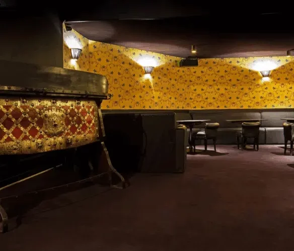 LE LOFT OHARA (75006) est un salon faiblement éclairé avec un papier peint jaune à motifs, une moquette sombre, des banquettes touffetées le long des murs, de petites tables rondes avec des chaises, et un objet orné partiellement visible sur la gauche.