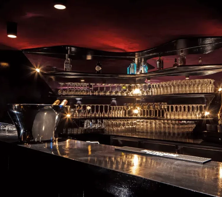 LE LOFT OHARA (75006) dispose d'un bar moderne, faiblement éclairé, avec un comptoir noir épuré, des verres à vin sur des étagères, des bouteilles d'alcool exposées et un seau à glace en argent contenant du champagne, le tout rehaussé par un éclairage chaleureux pour une ambiance chaleureuse.
