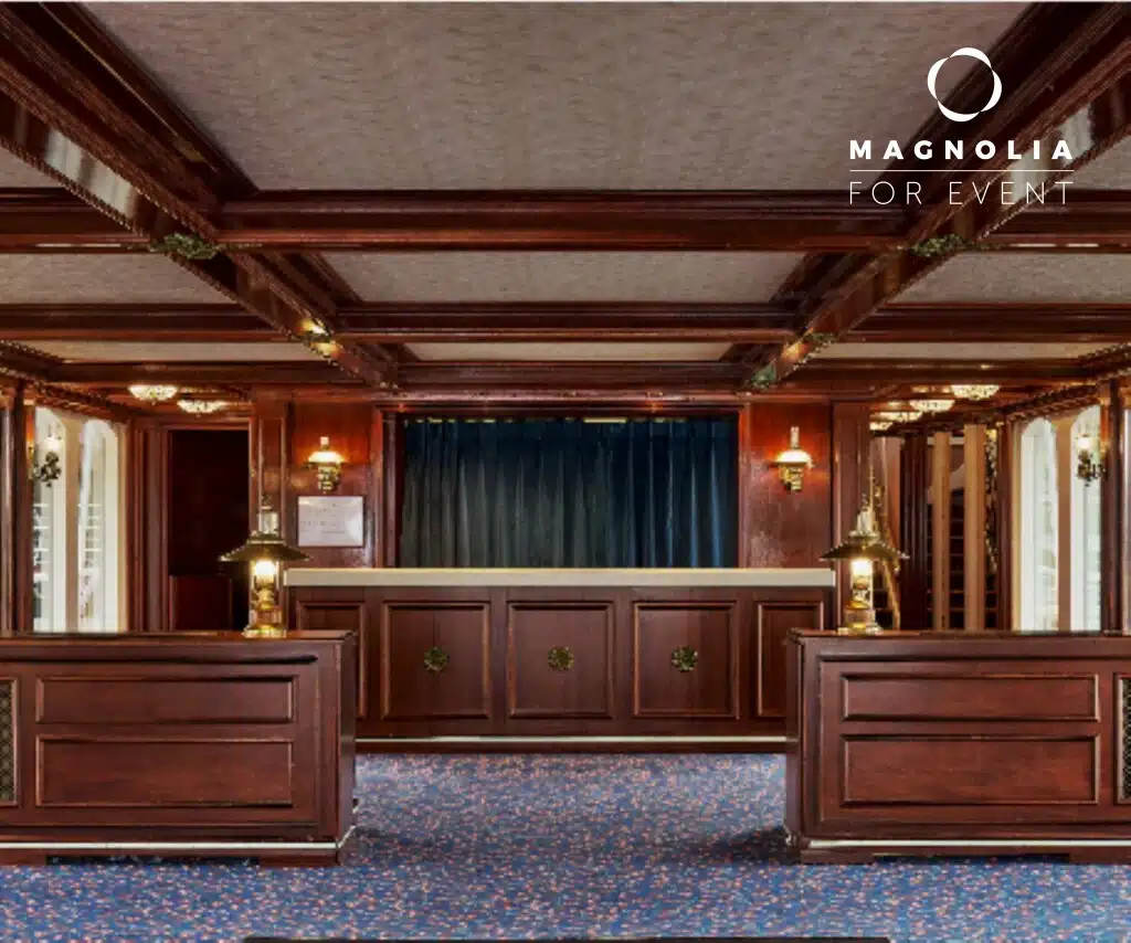 LE PETIT MISSISSIPI (75015) dispose d'une salle spacieuse et élégante avec des boiseries foncées, une moquette bleue, des lampes classiques en laiton et de grandes fenêtres. Le logo Magnolia For Event figure dans le coin supérieur droit.