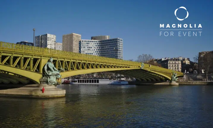 Un pont jaune avec des sculptures décoratives enjambe une rivière calme, entourée de bâtiments modernes et historiques sous un ciel clair. Le logo LE PETIT MISSISSIPI (75015) est visible dans le coin supérieur droit.