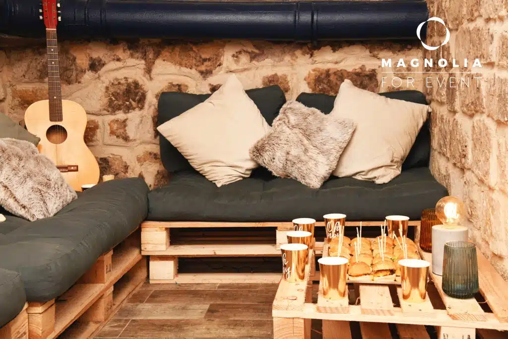 Coin salon confortable à LE PETIT LOFT FACTORY (75019) avec des canapés en palettes de bois, des coussins gris et en fourrure, une guitare, des snacks sur une table avec des tasses dorées, le tout contre un mur en pierre rustique. Logo Magnolia For Event dans le coin.