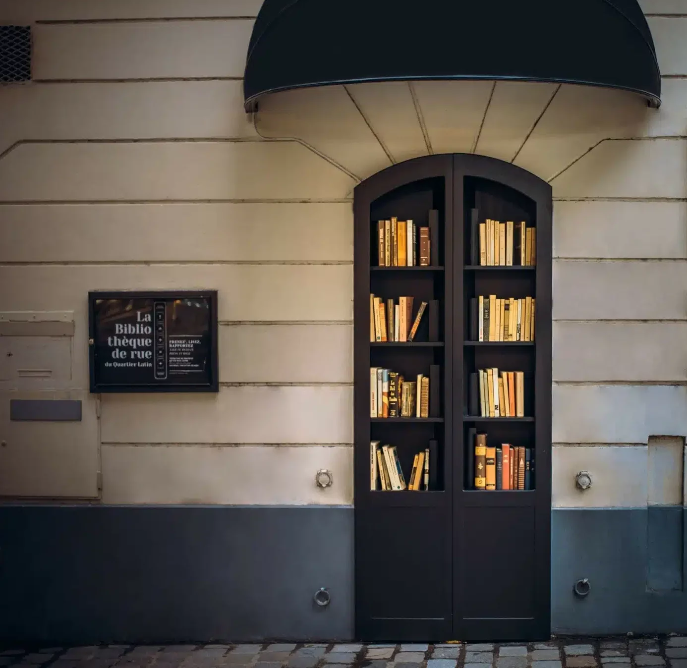 Une bibliothèque de rue encastrée dans un mur sous un auvent noir contient des livres. Sur la gauche, un panneau indique "LE LIEU SECRET (75005)". Le décor est un trottoir pavé.